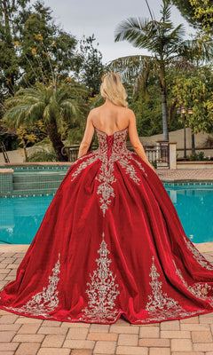 A woman wearing the Quinceanera Dress 1851 By Dancing Queen, featuring silver lace embroidery and a strapless design, stands by a pool with palm trees and patio furniture in the background, facing away from the camera.