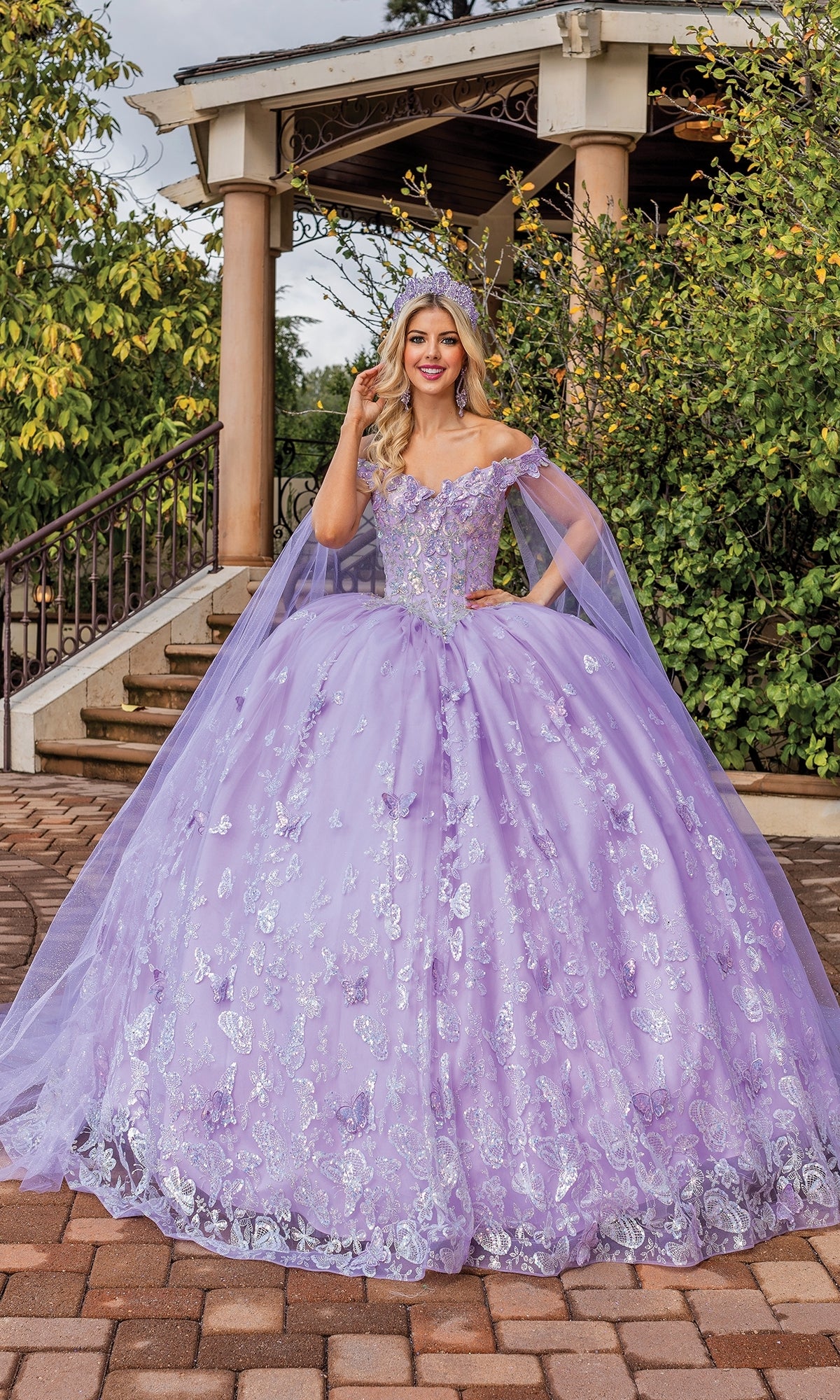 A young woman smiles at the camera outdoors in front of a gazebo, wearing Quinceanera Dress 1852 By Dancing Queen—a lilac off-shoulder ball gown with silver embroidery, draped cape sleeves, and a matching crown.