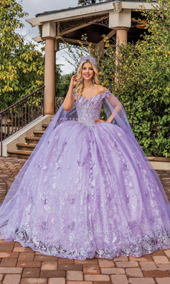 A young woman smiles at the camera outdoors in front of a gazebo, wearing Quinceanera Dress 1852 By Dancing Queen—a lilac off-shoulder ball gown with silver embroidery, draped cape sleeves, and a matching crown.