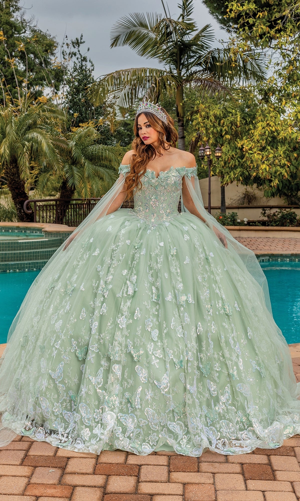 A woman in the Quinceanera Dress 1852 By Dancing Queen, an off-shoulder a-line ball gown with silver embroidery and a tiara, stands by a pool surrounded by palm trees.