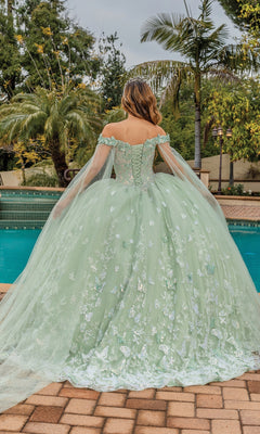 A woman wearing the Quinceanera Dress 1852 By Dancing Queen, featuring an a-line silhouette, draped cape sleeves, and floral details, stands by a pool with her back to the camera, surrounded by palm trees.