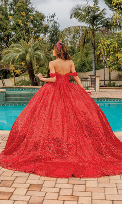 A woman wearing the Quinceanera Dress 1854 By Dancing Queen, a sparkling red A-line ball gown with a sweetheart neckline, stands by a pool outdoors with palm trees and greenery in the background, facing away.