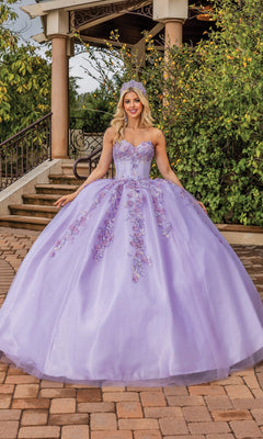 A woman poses outdoors on a brick path in front of greenery and a gazebo, wearing the Quinceanera Dress 1855 By Dancing Queen—a lilac gown with floral embellishments, strapless sweetheart neckline, and tiara that enhance her enchanting look.