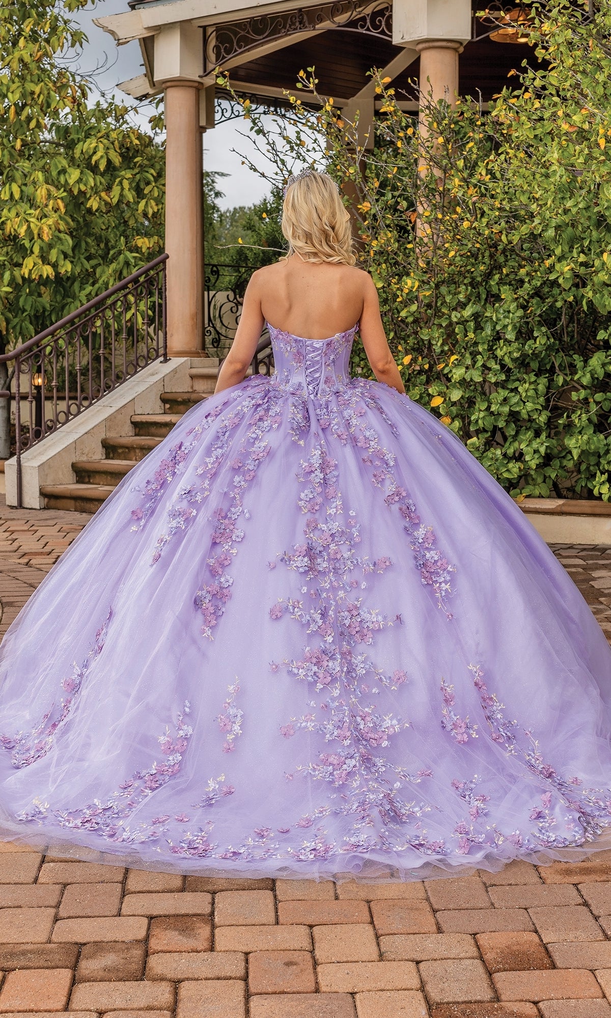 A person with blonde hair wears the Quinceanera Dress 1855 By Dancing Queen, a strapless lilac gown with a sweetheart neckline and floral appliqués, standing on a brick pathway near greenery and a gazebo.