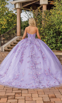 A person with blonde hair wears the Quinceanera Dress 1855 By Dancing Queen, a strapless lilac gown with a sweetheart neckline and floral appliqués, standing on a brick pathway near greenery and a gazebo.