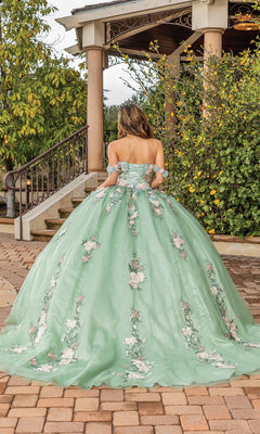 A woman stands outdoors near steps and greenery, facing away from the camera in Quinceanera Dress 1856 By Dancing Queen—an off-the-shoulder ball gown with floral details in soft sage green that beautifully complements the lush setting.