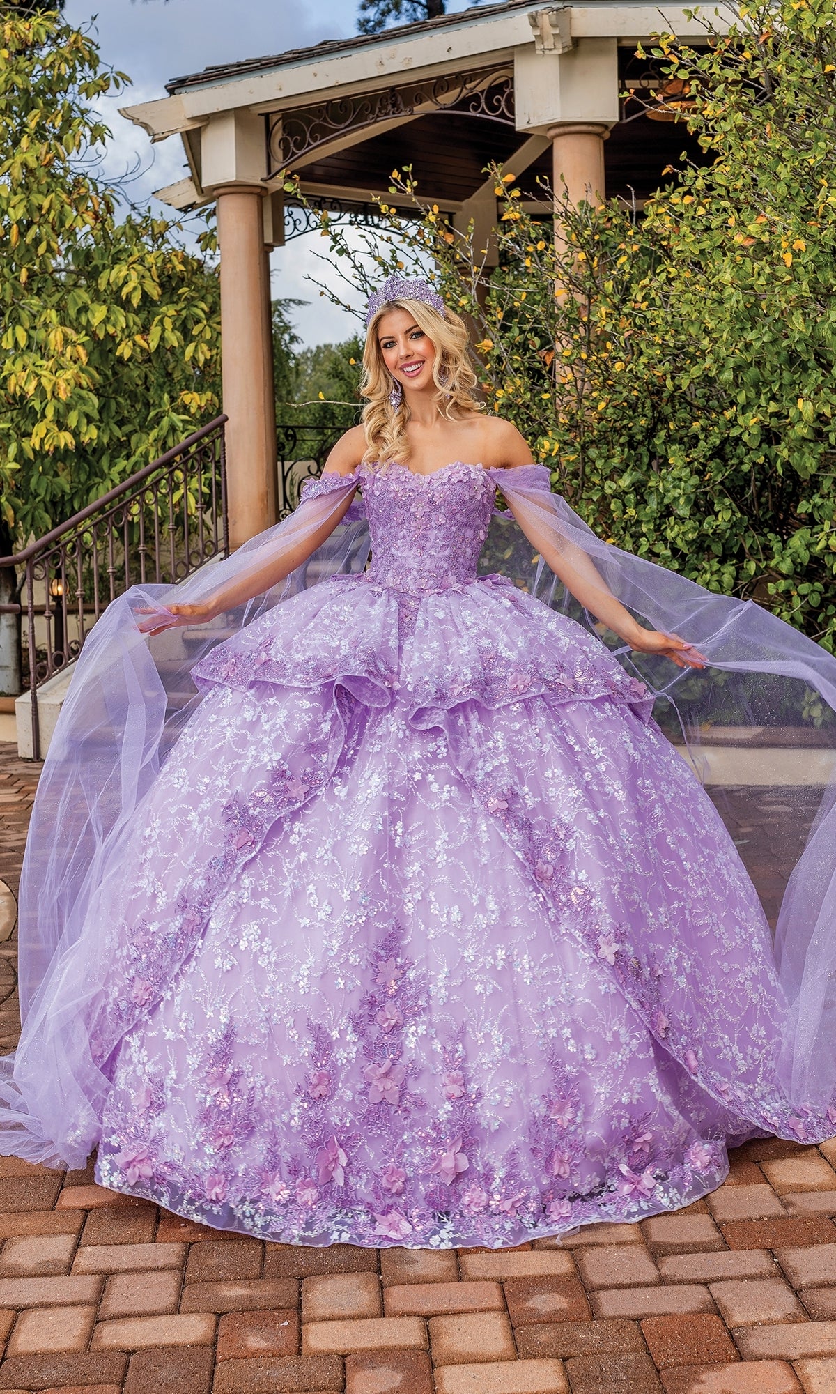A woman poses outdoors in front of greenery and a gazebo, wearing the Quinceanera Dress 1859 By Dancing Queen, a lilac purple floral-patterned tulle ball gown.