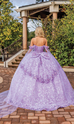 A woman in the Quinceanera Dress 1859 by Dancing Queen, featuring lilac purple fabric, off-shoulder cape sleeves, and a large bow, stands on a stone path near an outdoor structure surrounded by greenery.