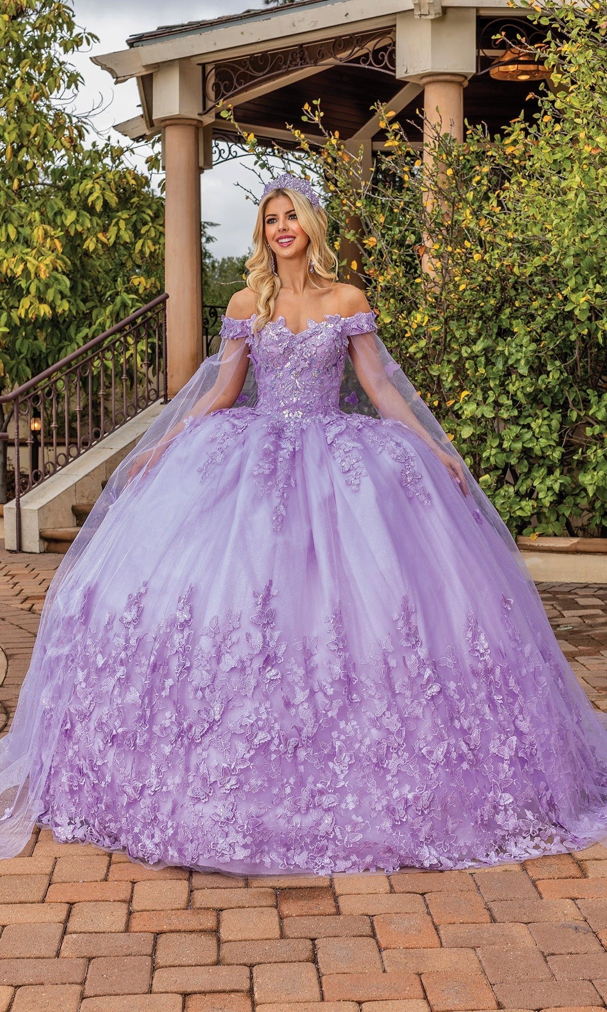 A woman stands outdoors smiling at the camera, wearing the Quinceanera Dress 1860 By Dancing Queen in lilac—a voluminous tulle ballgown with delicate floral appliqués and off-the-shoulder sleeves.