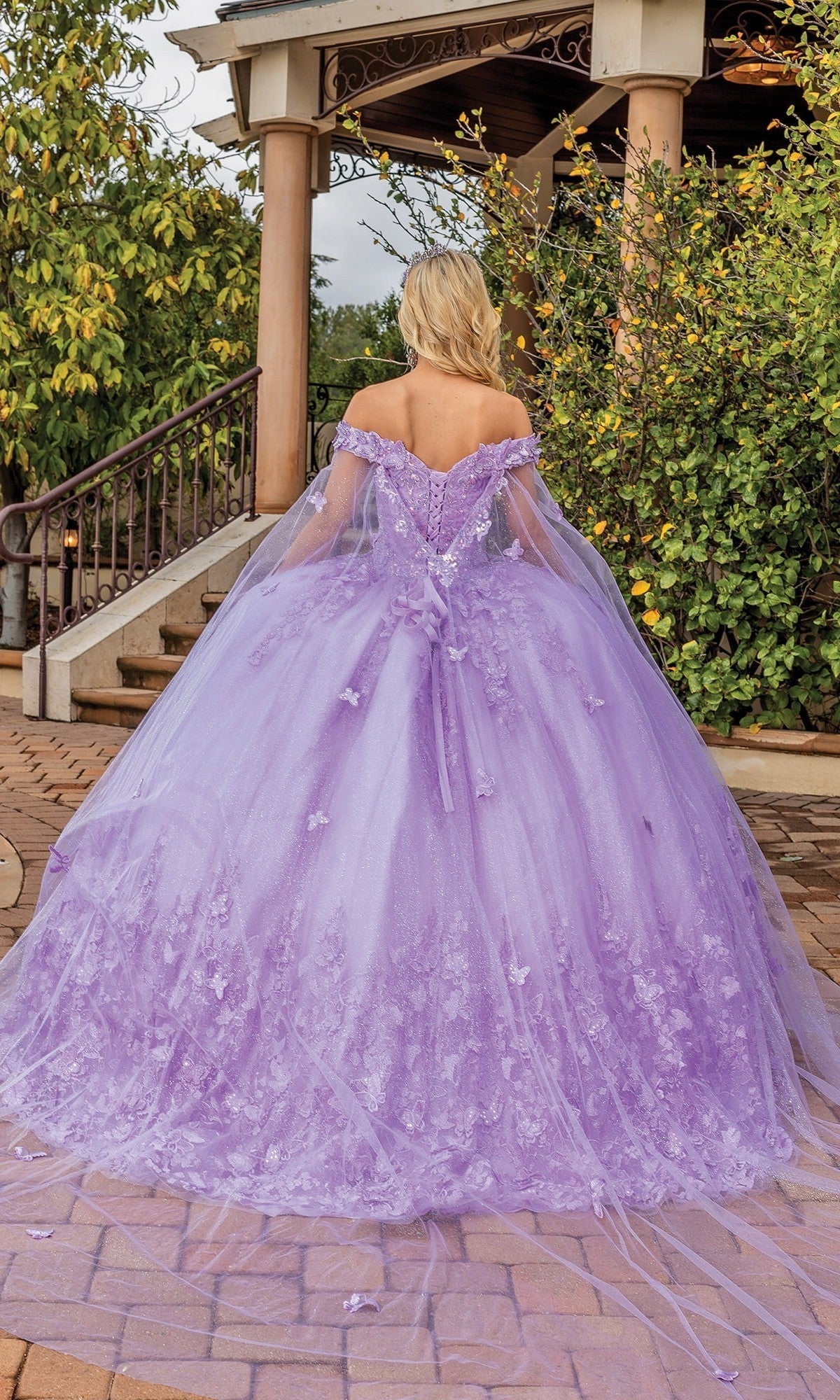 Outdoors near steps and greenery, a person faces away from the camera wearing the Quinceanera Dress 1860 By Dancing Queen—a lilac gown adorned with delicate floral appliques.