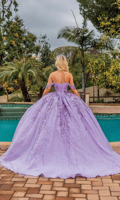 A woman in the Quinceanera Dress 1862 By Dancing Queen, a lilac off-the-shoulder ball gown, stands by a swimming pool with her back to the camera, surrounded by lush greenery and palm trees.