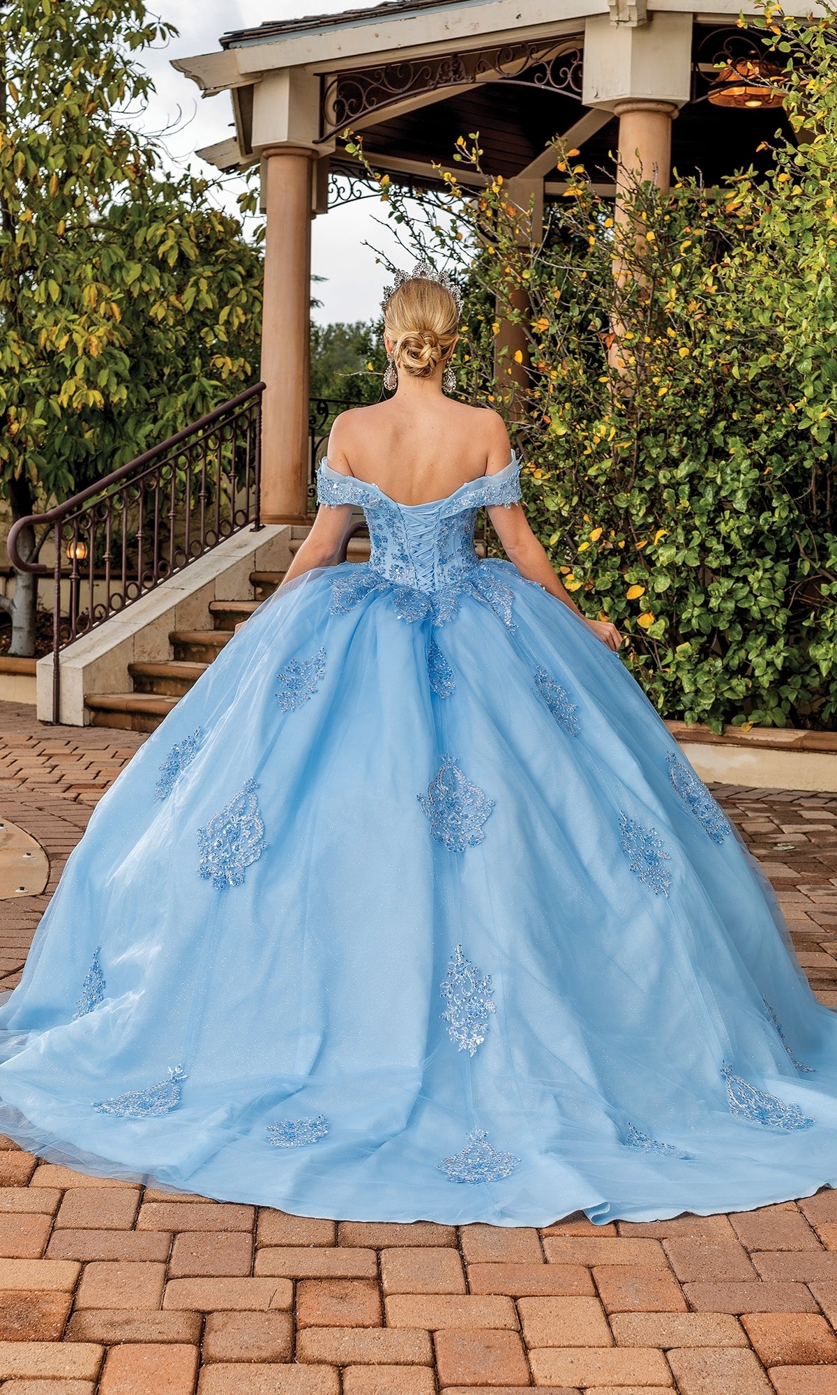 A person stands outdoors facing away from the camera in the Quinceanera Dress 1863 By Dancing Queen, an off-the-shoulder light blue ball gown with delicate lace applique and decorative embroidery.