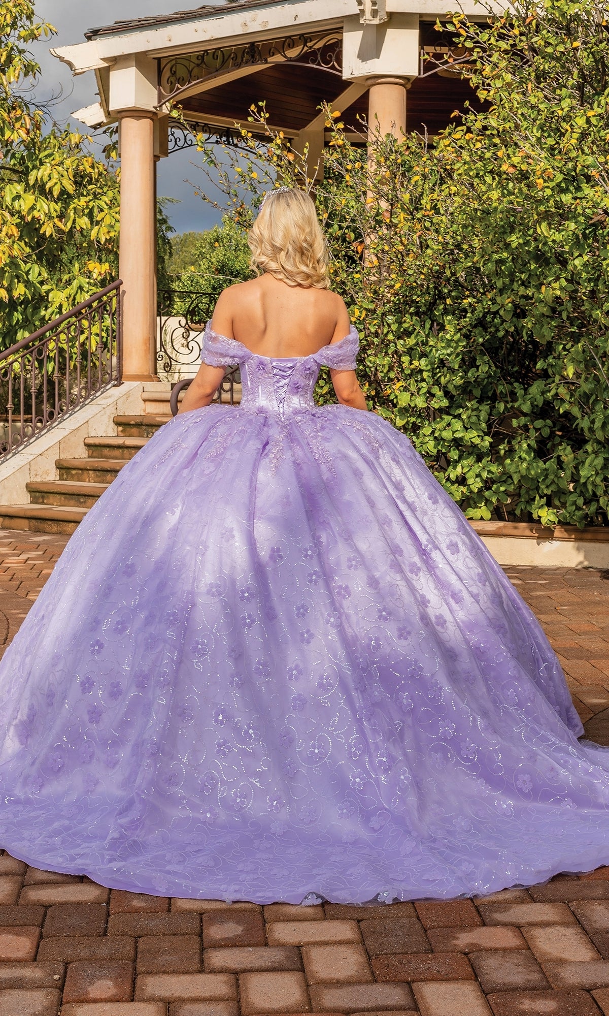 A blonde woman stands outside by gazebo steps, wearing the Quinceanera Dress 1864 By Dancing Queen—an off-shoulder, lilac-purple ball gown that flows elegantly around her.