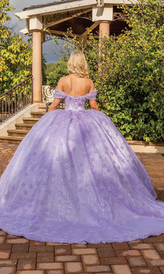 A blonde woman stands outside by gazebo steps, wearing the Quinceanera Dress 1864 By Dancing Queen—an off-shoulder, lilac-purple ball gown that flows elegantly around her.