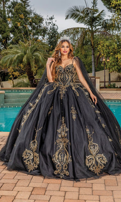 A woman wearing the Quinceanera Dress 1866 By Dancing Queen, featuring black fabric with gold embroidery and a sweetheart neckline, stands by a swimming pool amid palm trees and lush greenery.
