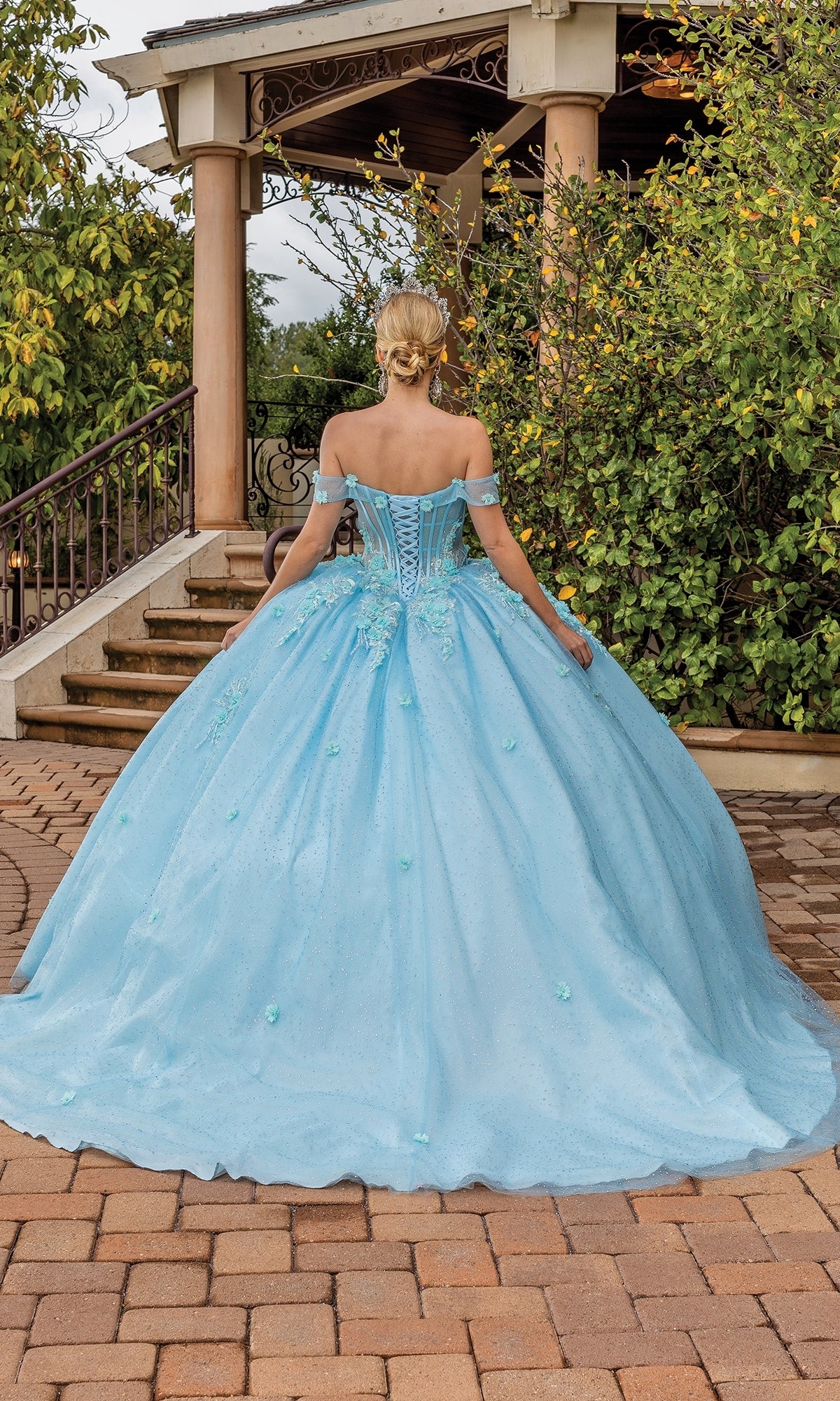 A woman wearing the Quinceanera Dress 1867 By Dancing Queen in aqua blue stands outdoors, facing away near stairs and greenery; the off-the-shoulder ball gown features a full skirt.