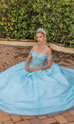 A woman in the Quinceanera Dress 1867 By Dancing Queen, an aqua blue off-the-shoulder ball gown, sits outdoors on a brick surface with lush green bushes behind her, wearing a sparkling tiara.