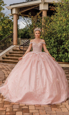 A woman wearing the Quinceanera Dress 1868 by Dancing Queen—pink, off-the-shoulder with a full skirt—stands outdoors on a stone path in front of a gazebo, crowned and radiant.