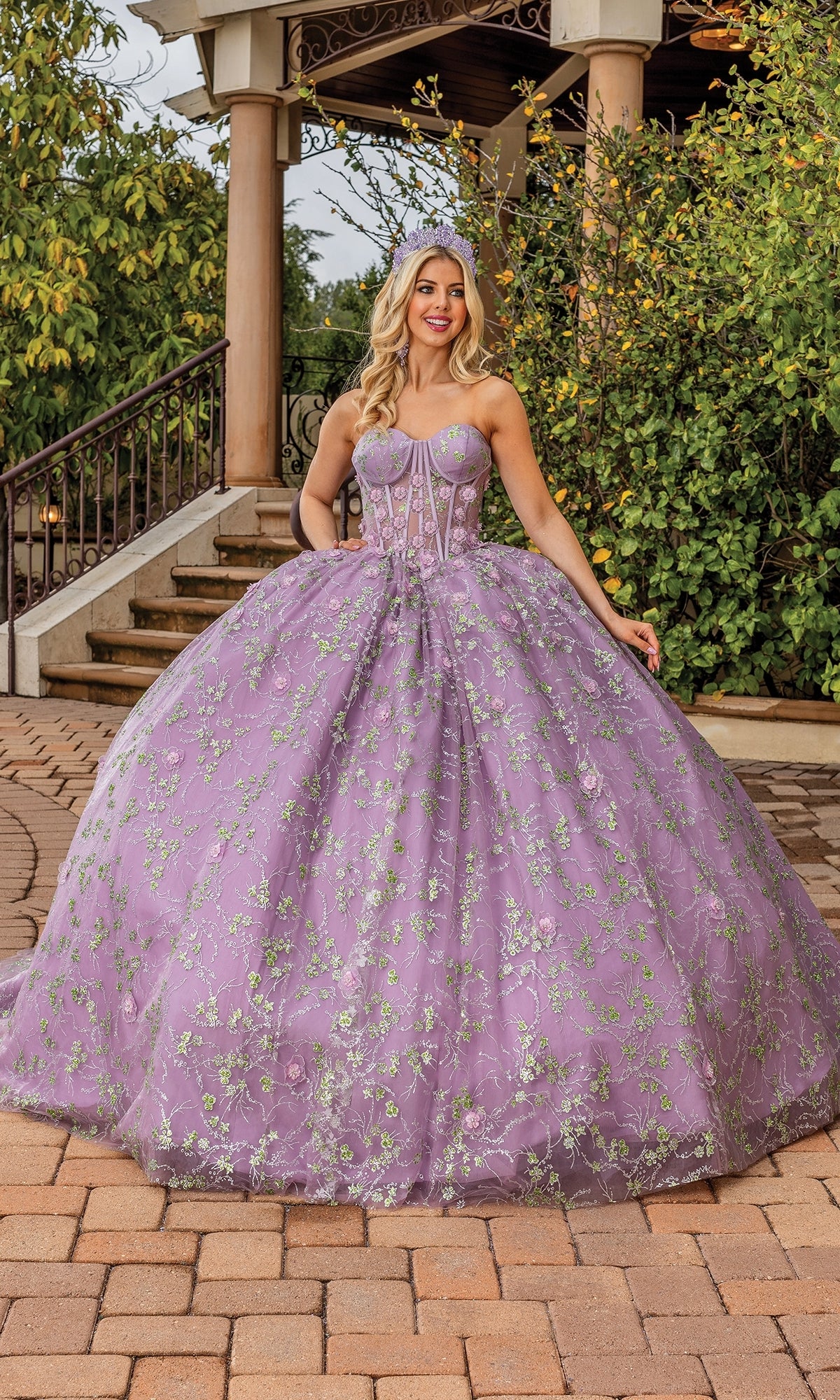 A woman poses outdoors in front of stairs and greenery, wearing the Quinceanera Dress 1872 by Dancing Queen—a dusty lilac gown with floral embroidery and a strapless sweetheart neckline.