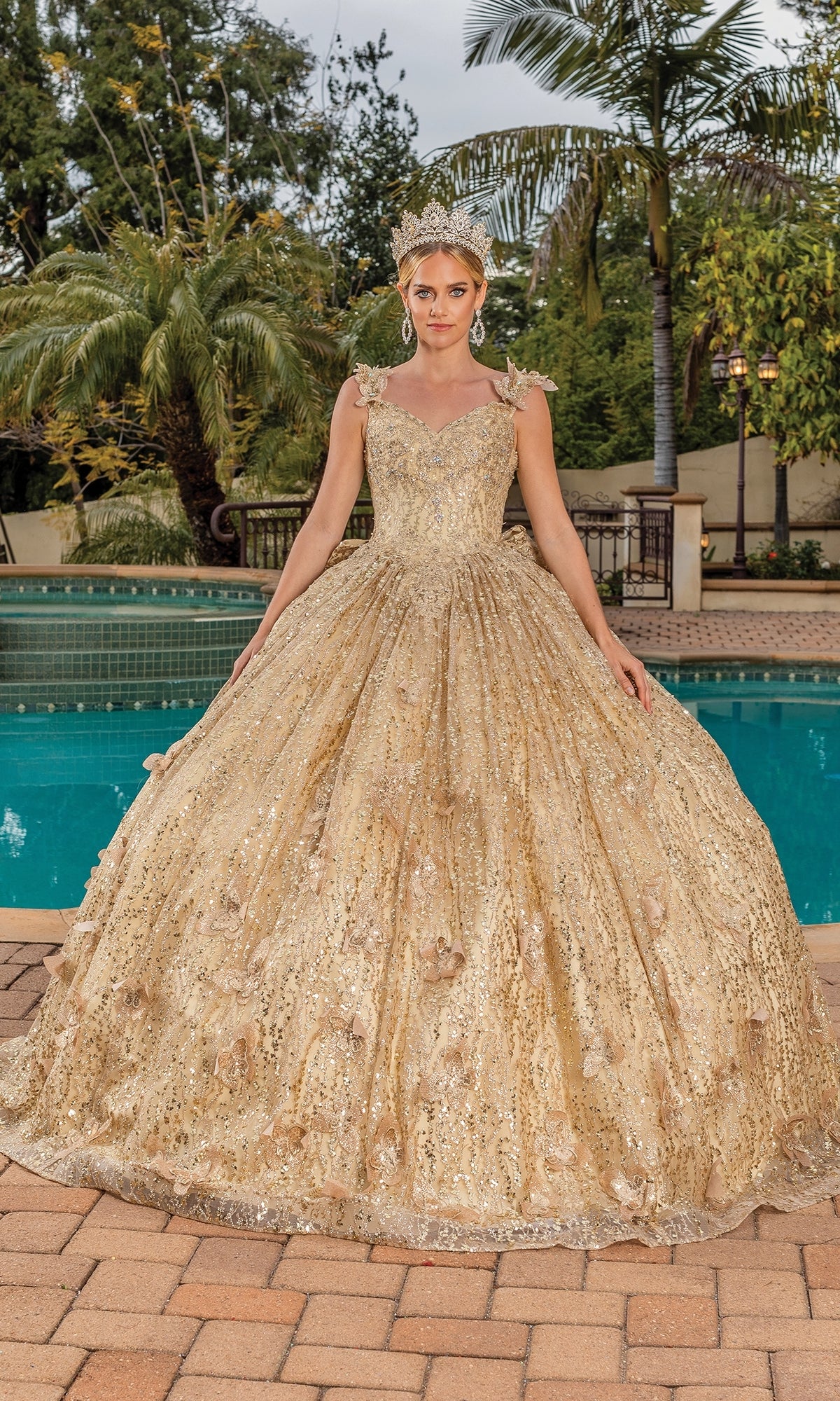 A woman poses by a pool in the Quinceanera Dress 1877 By Dancing Queen, featuring a gold full skirt, sequin details, removable puff sleeves, and a matching tiara. Palm trees and lush greenery create an elegant backdrop.