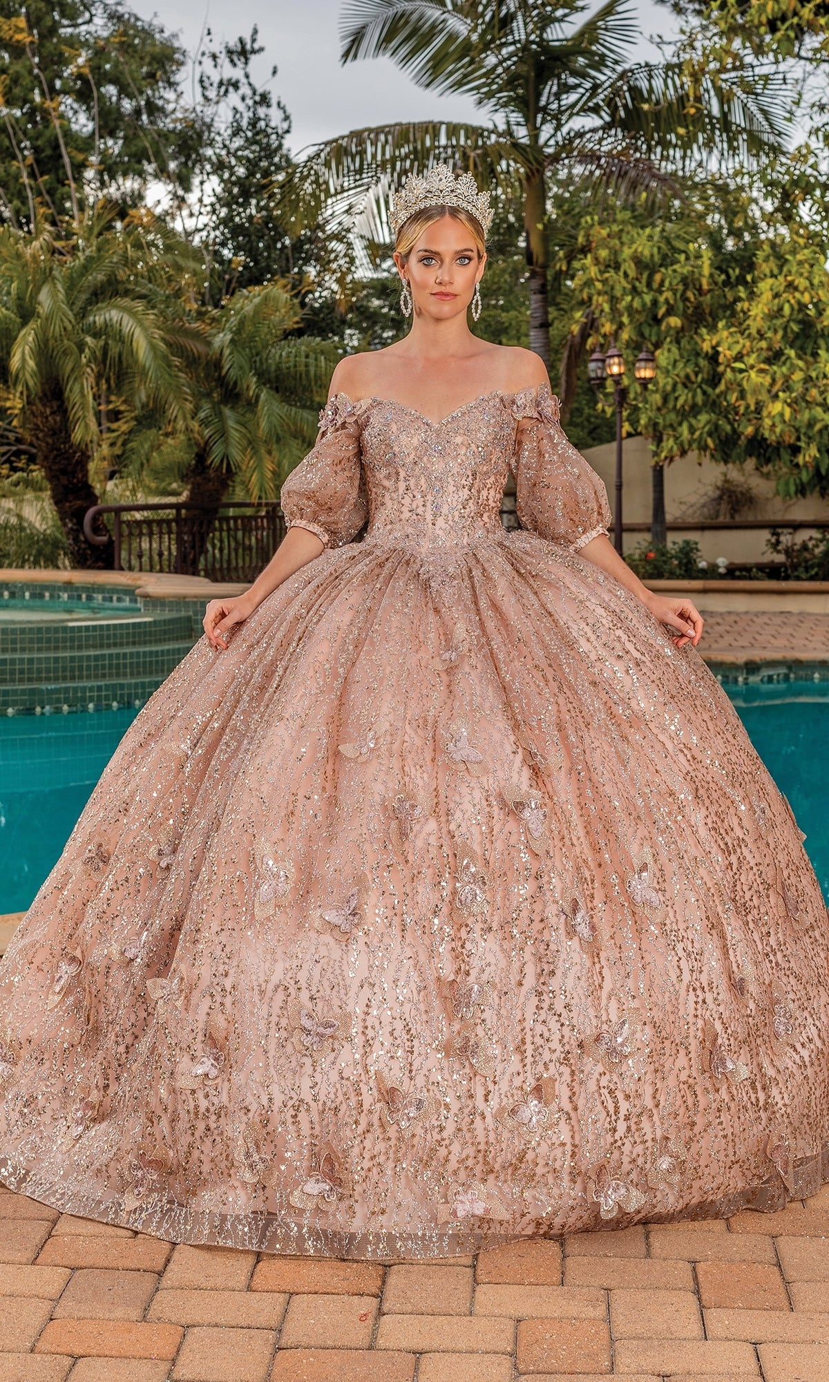 A woman poses by a pool in the Quinceanera Dress 1877 By Dancing Queen, an ornate gold a-line ball gown with removable puff sleeves and sparkling embellishments, surrounded by lush greenery.