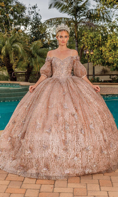 A woman poses by a pool in the Quinceanera Dress 1877 By Dancing Queen, an ornate gold a-line ball gown with removable puff sleeves and sparkling embellishments, surrounded by lush greenery.