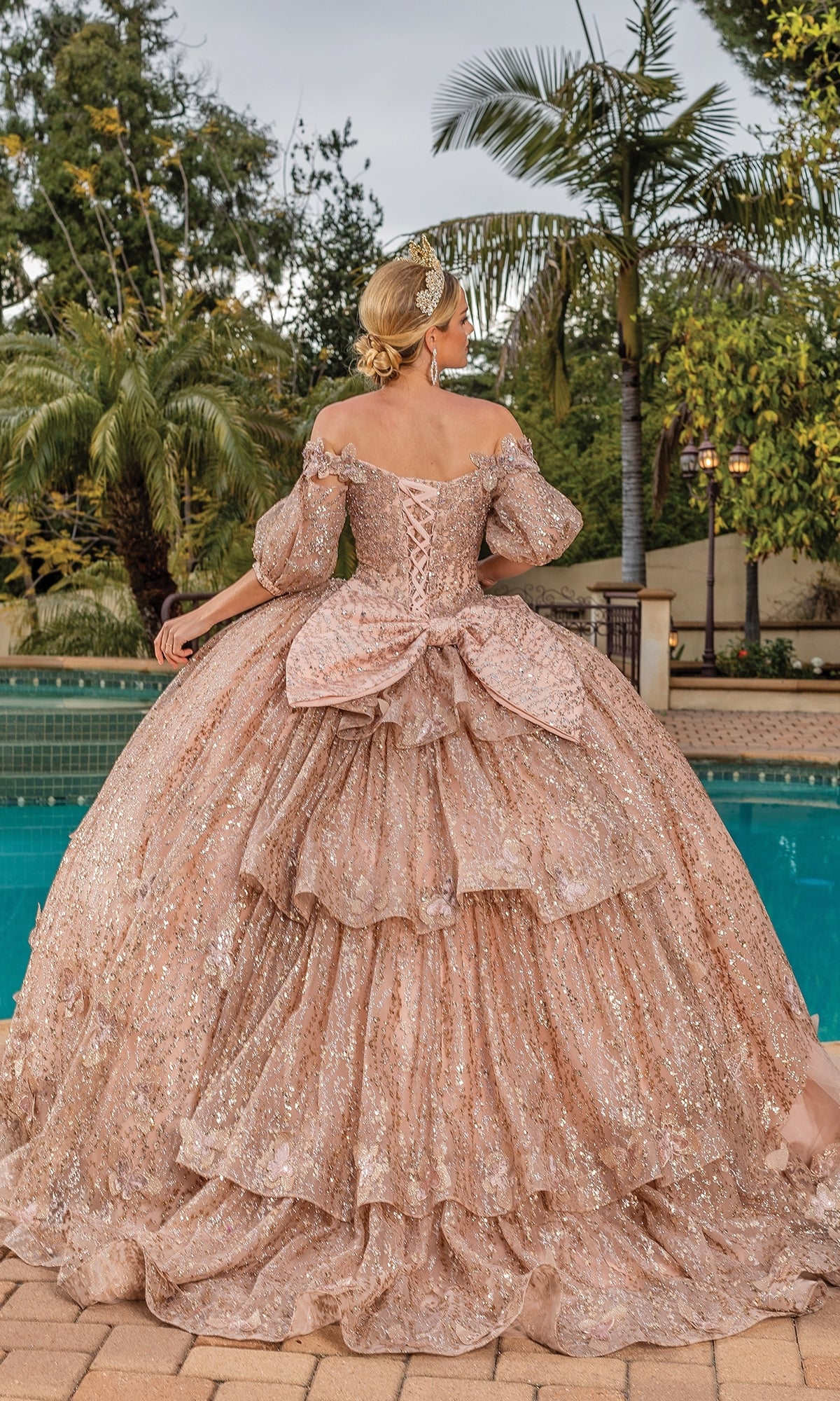 A woman stands by a pool surrounded by palm trees, wearing the Quinceanera Dress 1877 By Dancing Queen—a sparkling off-the-shoulder gold gown with a large bow and removable puff sleeves—facing away.