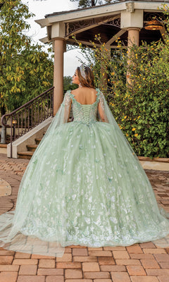 A person wearing the Quinceanera Dress 1878 By Dancing Queen—a light green, embroidered ball gown with long sheer sleeves and a sweetheart neckline—stands facing away from the camera before a gazebo surrounded by greenery.