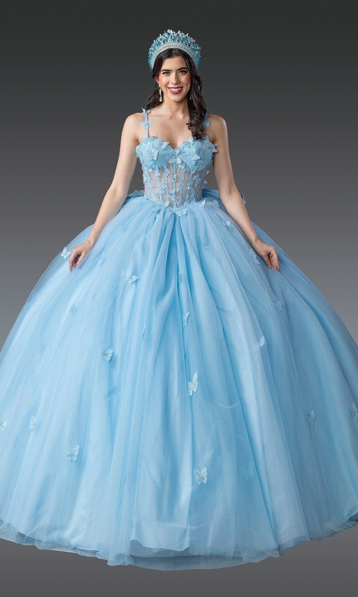 A woman stands against a gray background wearing the Quinceanera Dress 1879 by Dancing Queen in Bahama Blue, a ball gown adorned with floral and butterfly details, featuring a sweetheart neckline and a matching crown.