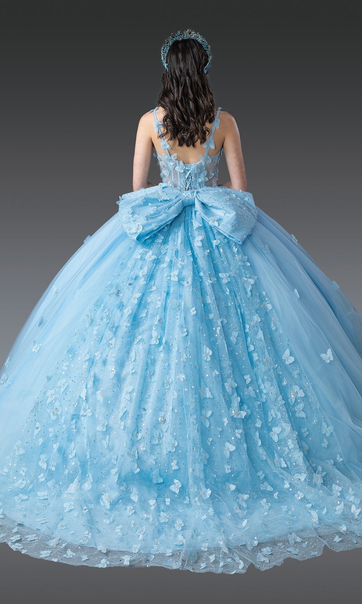 A woman is shown from behind against a plain gray background, wearing the Quinceanera Dress 1879 by Dancing Queen—a pastel ball gown adorned with butterfly appliqués, featuring a large back bow and a matching headpiece.