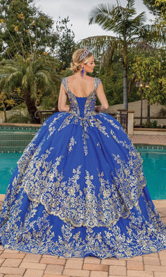 A woman wearing the Quinceanera Dress 1881 By Dancing Queen in royal blue with gold lace applique stands by a pool, facing away, surrounded by palm trees and lush greenery.