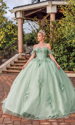 A woman in the Quinceanera Dress 1883 By Dancing Queen—a sage ball gown with a sweetheart neckline—wears a sparkling tiara as she stands on a brick path before a gazebo surrounded by greenery.