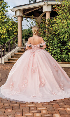 A person in the Quinceanera Dress 1883 by Dancing Queen, an off-the-shoulder pink ball gown with floral details, stands outdoors near stairs and a gazebo surrounded by greenery.