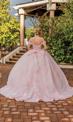 A woman in the Quinceanera Dress 1888 By Dancing Queen—a pink off-the-shoulder A-line ball gown with floral details and a large bow—stands outdoors facing a staircase surrounded by lush greenery.