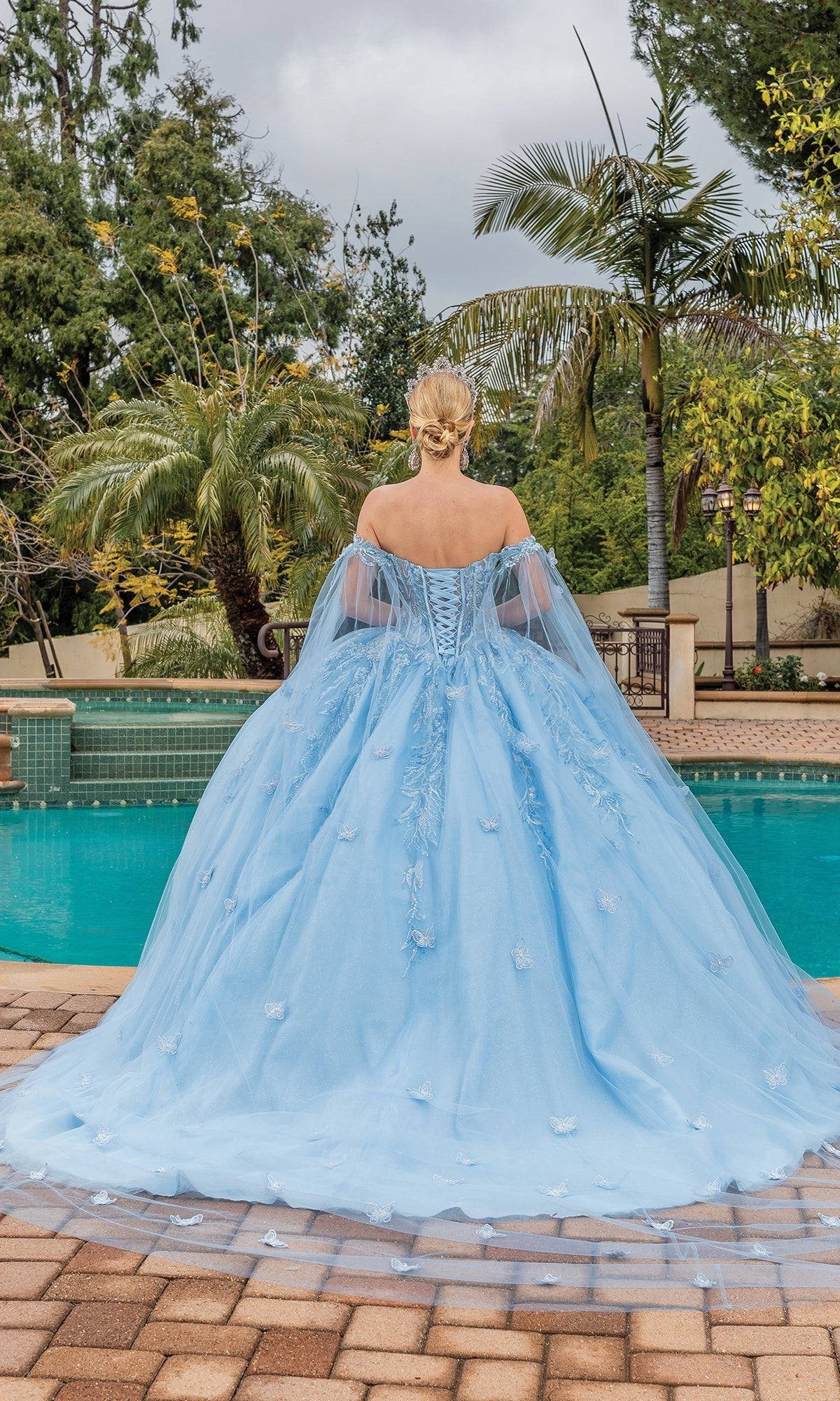 A woman stands by a pool, facing away in the Quinceanera Dress 1889 By Dancing Queen—a light blue, off-the-shoulder ball gown with a full skirt—surrounded by lush greenery and palm trees.