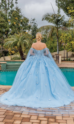 A woman stands by a pool, facing away in the Quinceanera Dress 1889 By Dancing Queen—a light blue, off-the-shoulder ball gown with a full skirt—surrounded by lush greenery and palm trees.