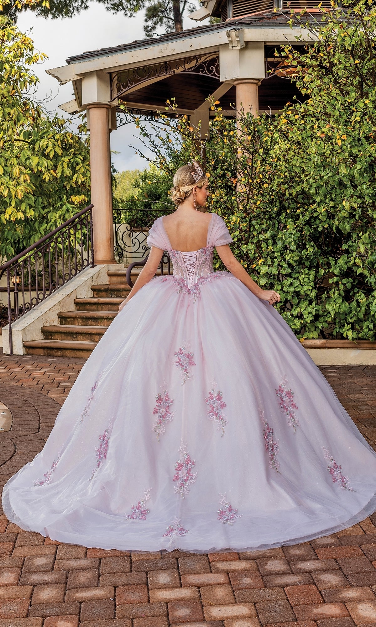 A woman in the Quinceanera Dress 1892 By Dancing Queen, a light purple a-line ball gown with floral details and a sweetheart neckline, stands on a stone path near a gazebo surrounded by greenery, facing away from the camera.