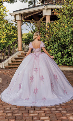 A woman in the Quinceanera Dress 1892 By Dancing Queen, a light purple a-line ball gown with floral details and a sweetheart neckline, stands on a stone path near a gazebo surrounded by greenery, facing away from the camera.