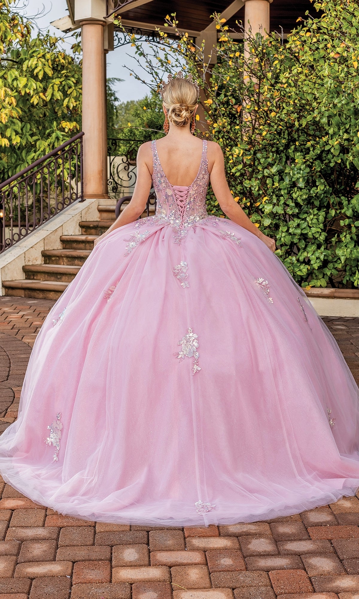 Someone stands outdoors facing away from the camera, wearing the Quinceanera Dress 1896 By Dancing Queen—a blush pink gown with floral embellishments, lace embroidery, and a lace-up back.