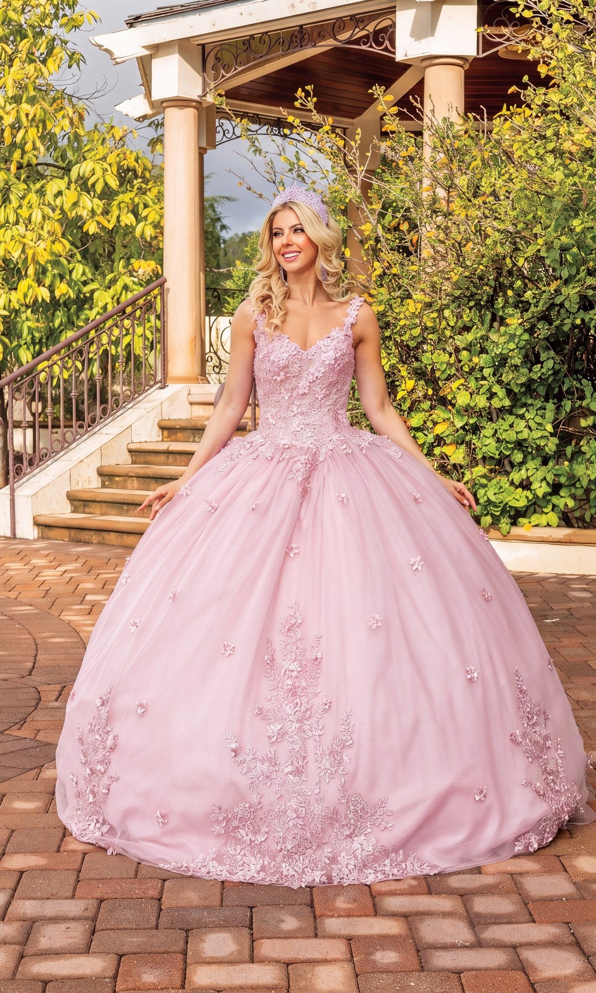 Wearing the Quinceanera Dress 1898 By Dancing Queen in blush, a woman stands outside on a stone patio near greenery and a gazebo; the gown features an elegant a-line silhouette with delicate floral embellishments and a graceful v-neckline.