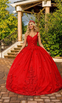 A woman wearing the Quinceanera Dress 1898 by Dancing Queen in red, featuring an a-line silhouette with intricate floral embellishments and a v-neckline, stands outdoors on a brick path near a gazebo and greenery, smiling and looking to the side.