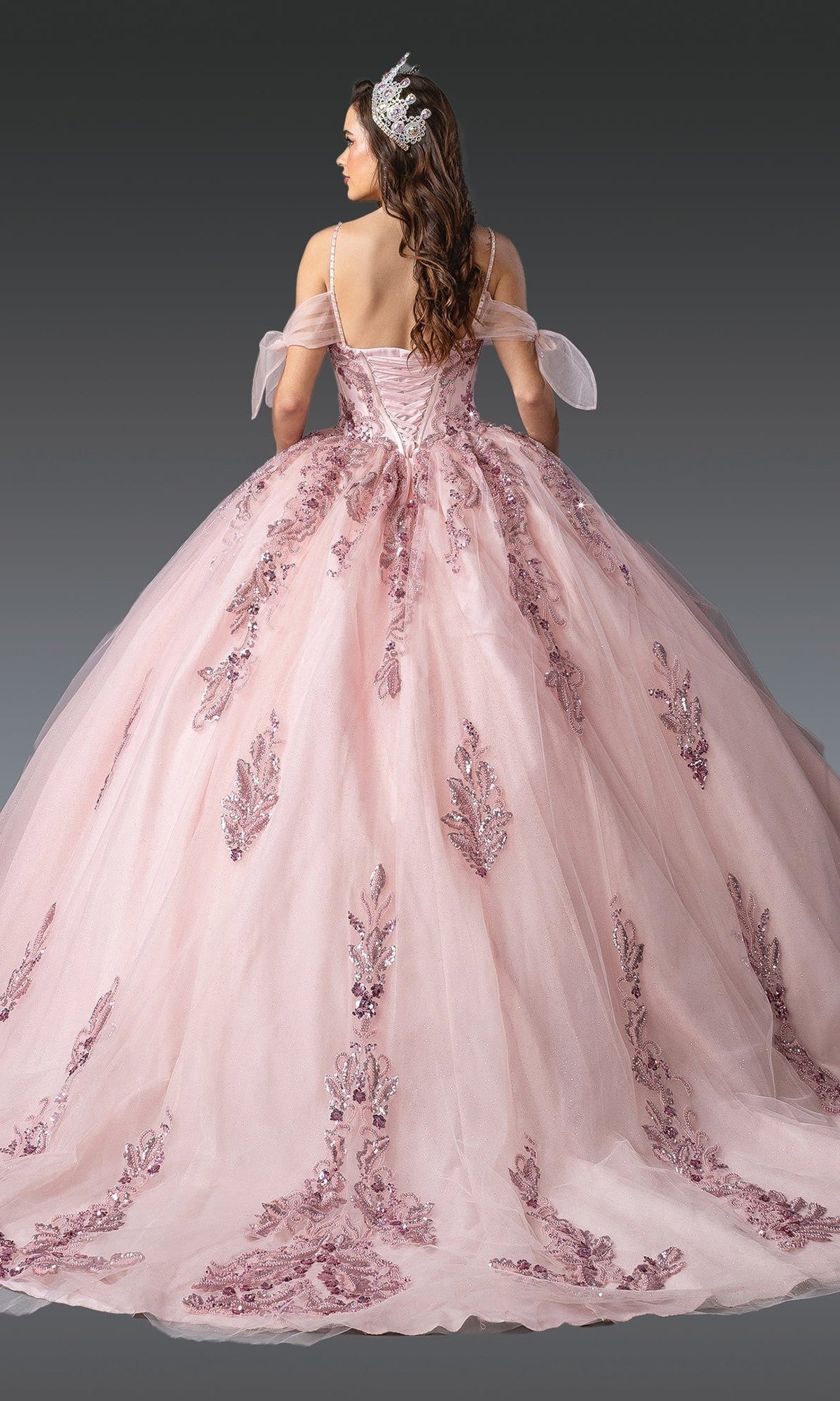 A woman faces away on a gray background, wearing the Quinceanera Dress 1902 by Dancing Queen—a pink tulle ballgown featuring off-the-shoulder sleeves and an embellished bodice adorned with sequin accents.