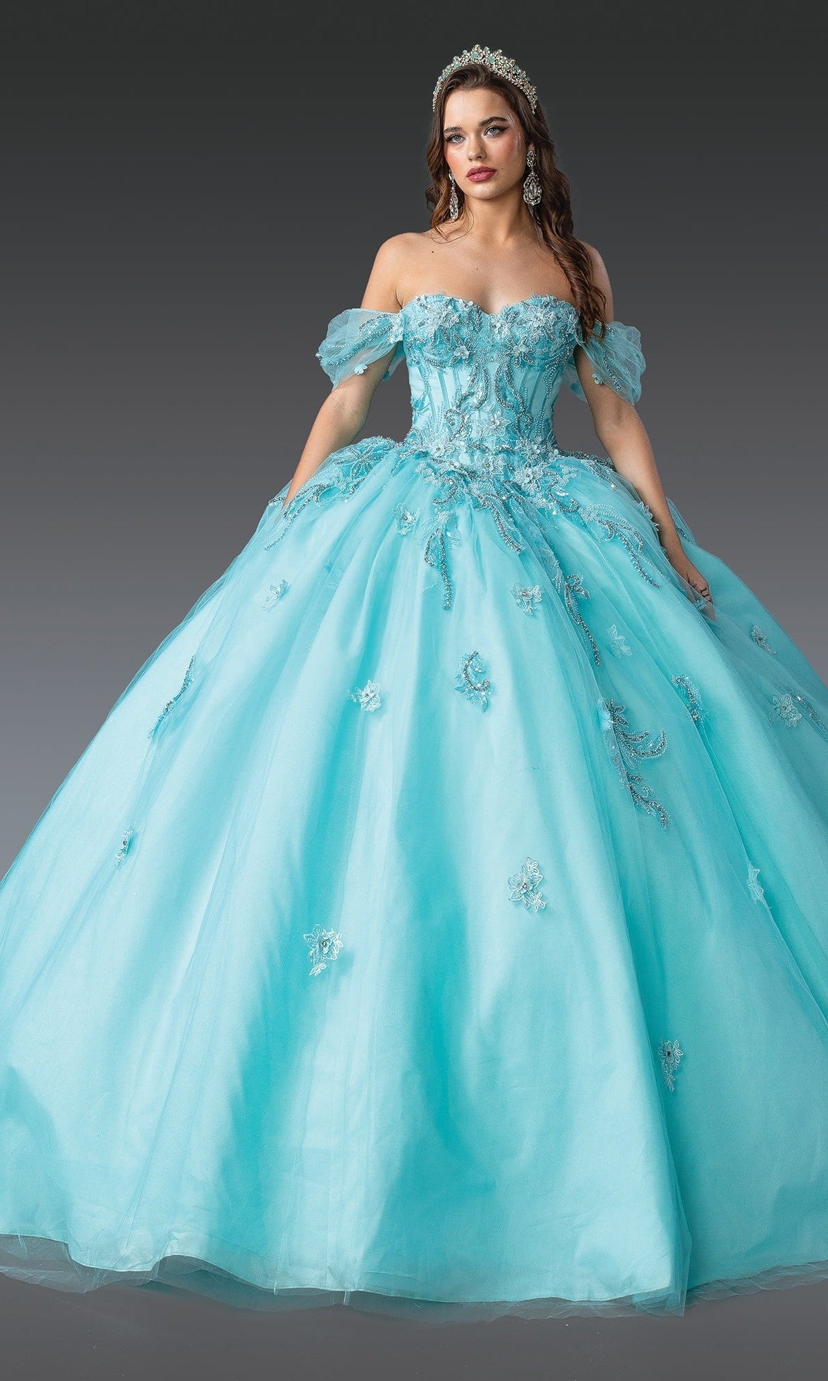 A woman in the Quinceanera Dress 1903 By Dancing Queen, an aqua off-the-shoulder ball gown with floral embellishments, a tiara, a fitted corset bodice, and an elegant ballgown skirt, standing against a gray background.