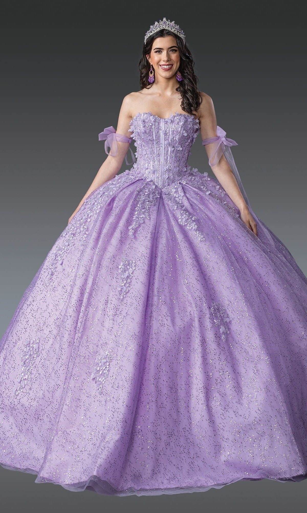 A person wearing the Quinceanera Dress 1905 by Dancing Queen in lilac, featuring a tiara and a glitter ball gown with a corset bodice and sparkling lavender floral embellishments, poses against a plain gray background.