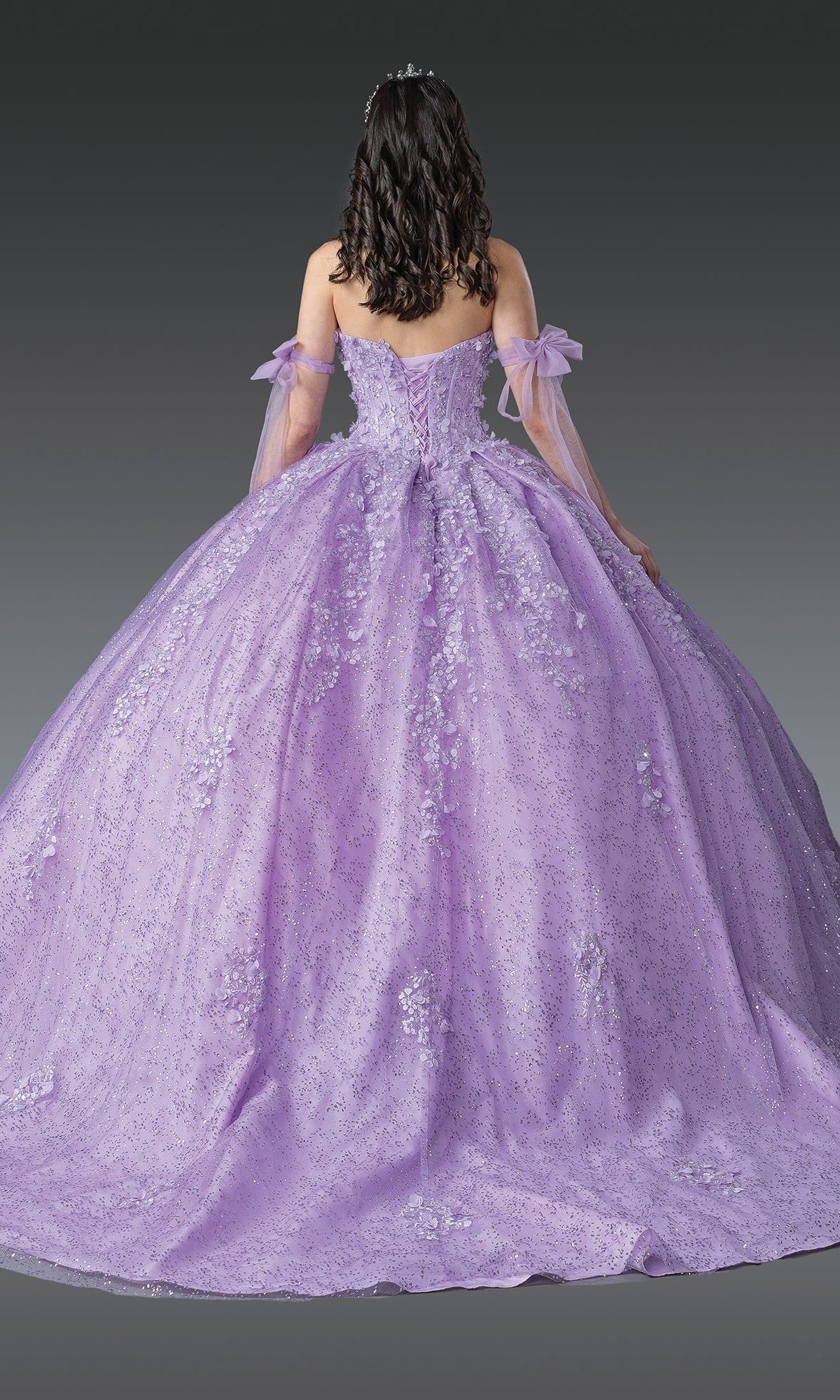 A person with curly dark hair is shown from the back against a plain gray background, wearing the Quinceanera Dress 1905 by Dancing Queen—a sparkling lilac gown featuring off-shoulder tulle sleeves and a fitted corset bodice.