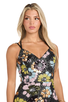 A woman with long blonde hair wears the Floral-Sequin Long Black Prom Dress: Morgan 19051, a black gown decorated with large, colorful floral sequins. She gazes slightly left with a neutral expression.