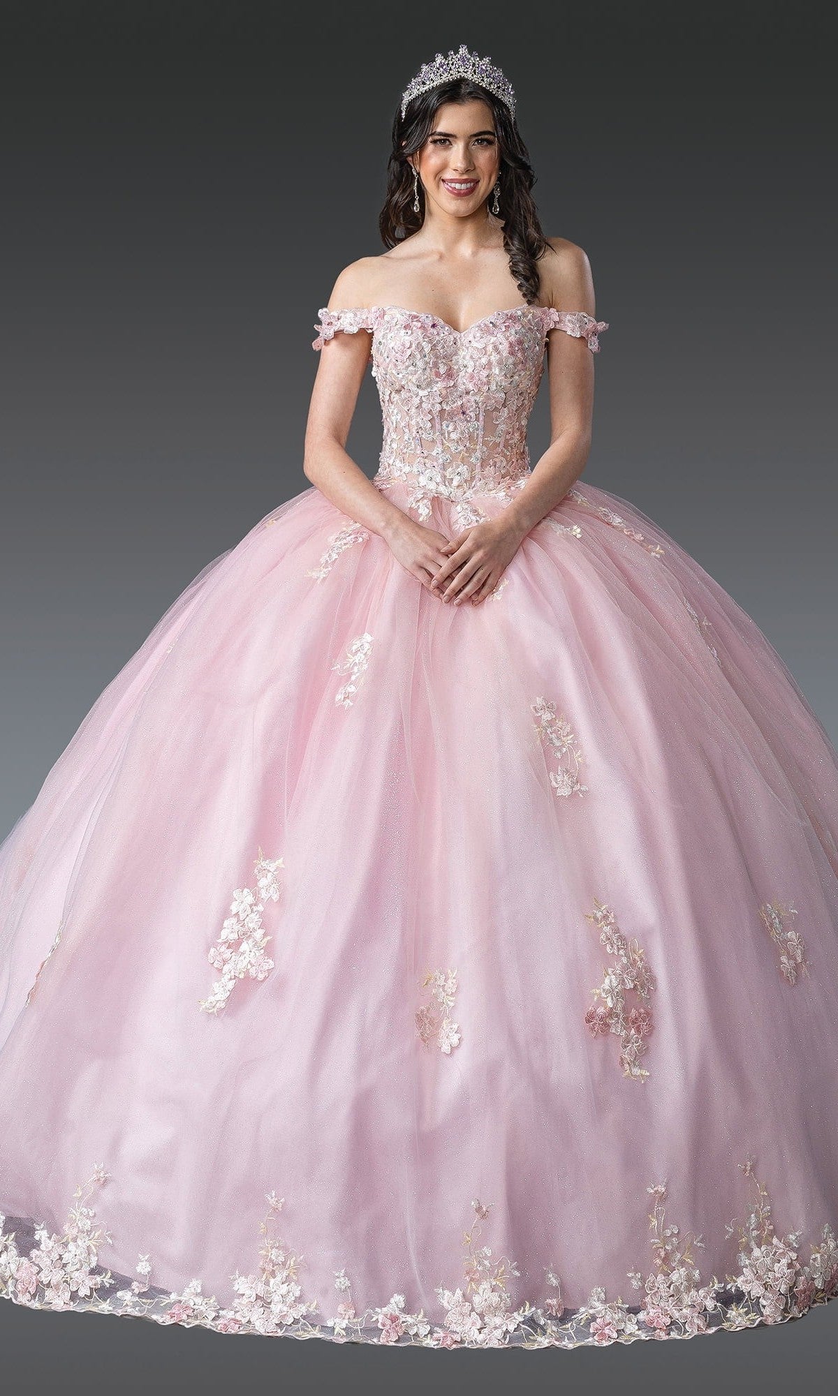 A woman wearing the Quinceanera Dress 1906 By Dancing Queen in blush, featuring an off-the-shoulder design with 3D floral appliques and a tiara, stands against a plain gray background.