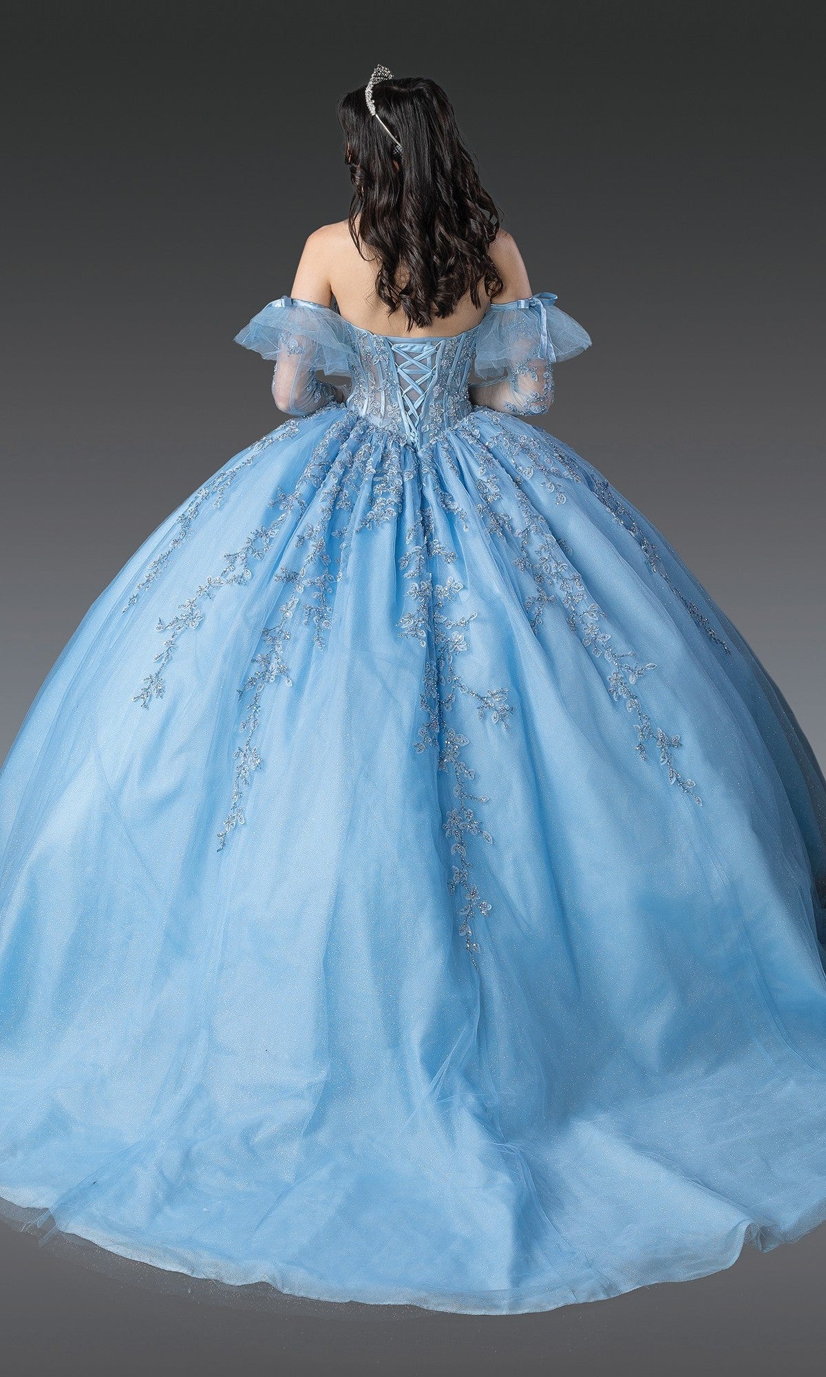 A person with long dark hair is photographed from the back, wearing the Quinceanera Dress 1910 By Dancing Queen—an off-shoulder gown highlighted by a blue ball gown skirt, silver embroidery, and an embellished corset bodice that adds an elegant touch.