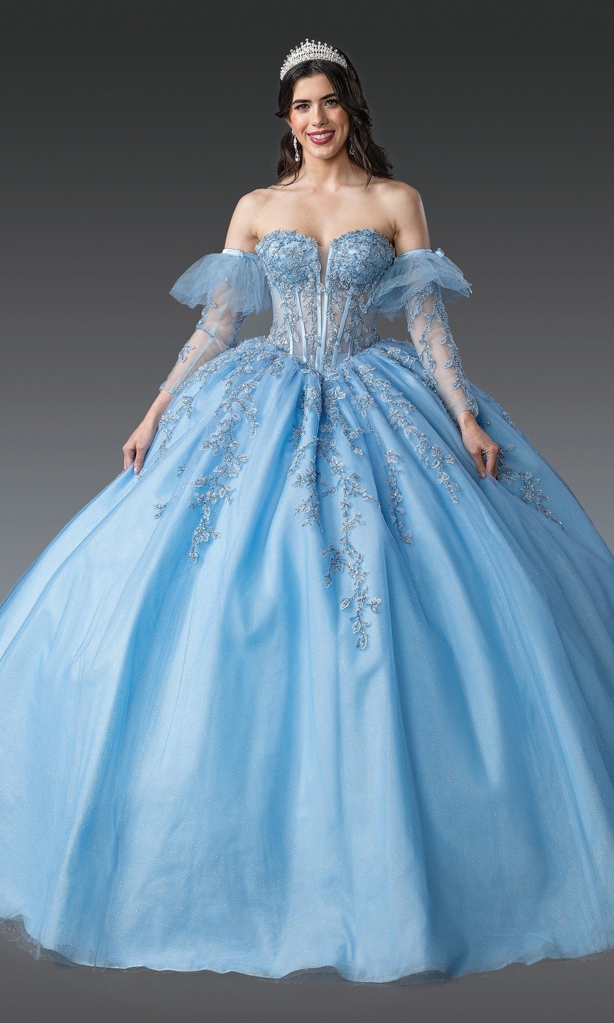 A woman wearing the Quinceanera Dress 1910 by Dancing Queen in Bahama Blue, featuring a silver-embroidered corset bodice, off-the-shoulder tulle sleeves, and a tiara, stands against a gray background.