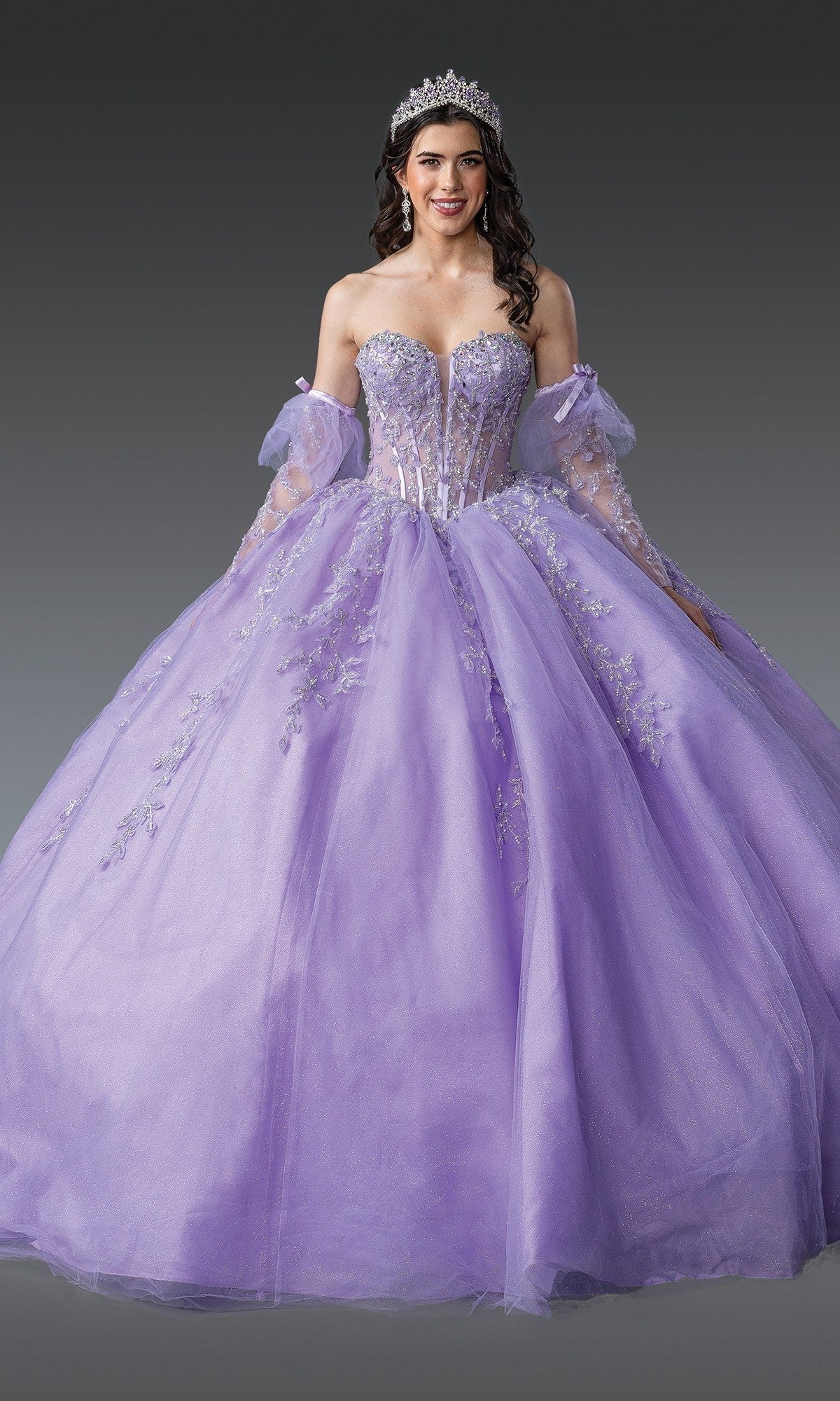 A woman wearing a silver tiara and the Quinceanera Dress 1910 By Dancing Queen in lilac, featuring an off-shoulder design, an embellished corset bodice, and floral embroidery on the ball gown skirt, stands against a plain gray background smiling at the camera.